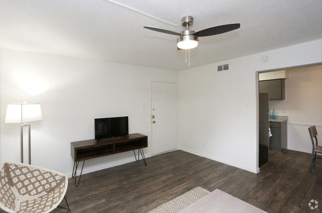 Living room with TV on a dark wooden stand, light-colored walls, and a view into the kitchen.
