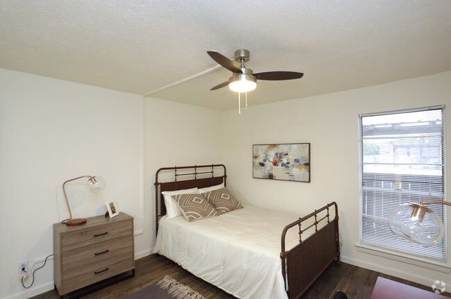 Bedroom with a brown metal bed, nightstand, and window with blinds. Ceiling fan hangs overhead.