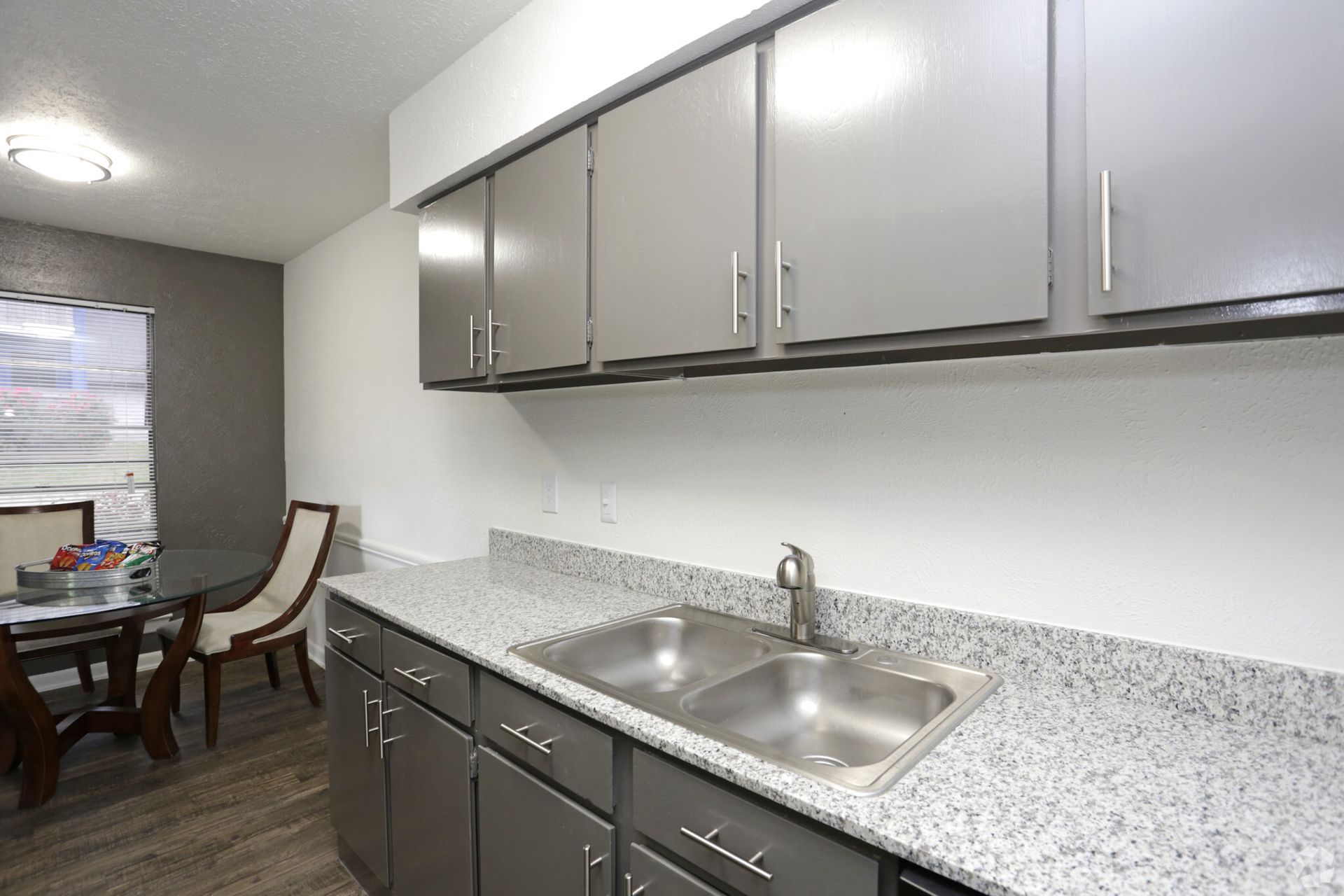 Kitchen with gray cabinets, a double sink, granite countertops, and a dining table by a window.
