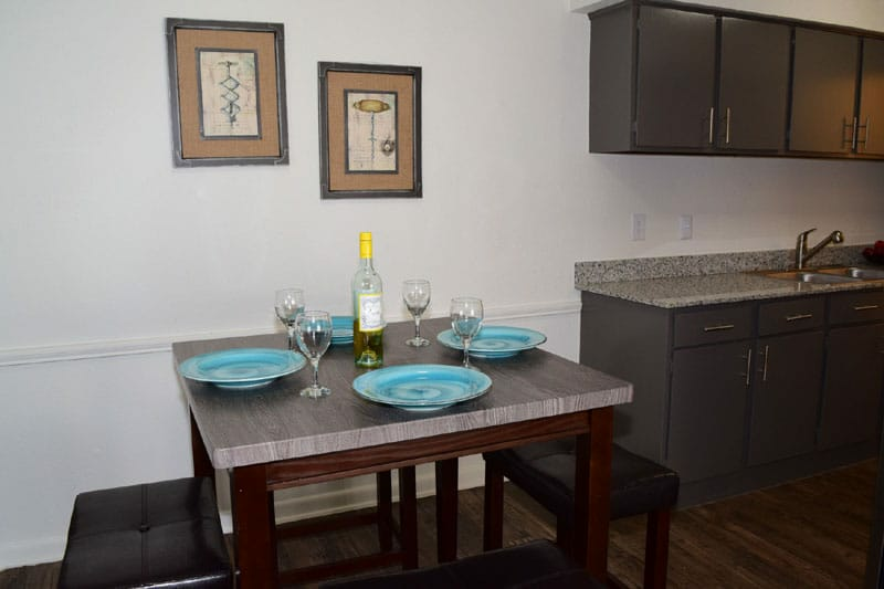 Dining area with a square table set for four, next to a kitchen with gray cabinets.