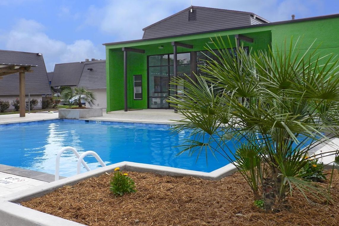 Pool with palm trees, bright green building, and dark roof under a blue sky.