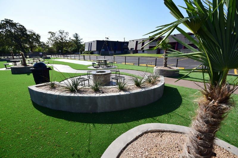 Park area with fire pit, grill, and seating on artificial turf, with buildings in the background.