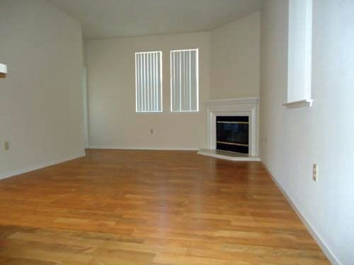 Empty living room with wood floor, white walls, two windows, and a fireplace.