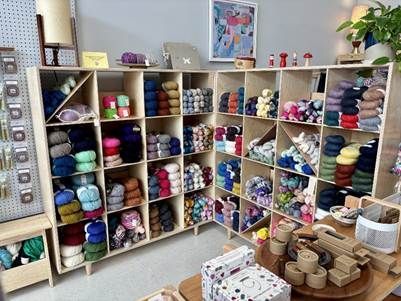 A craft store shelf filled with organized, colorful skeins of yarn, with art and supplies on a desk in the foreground.