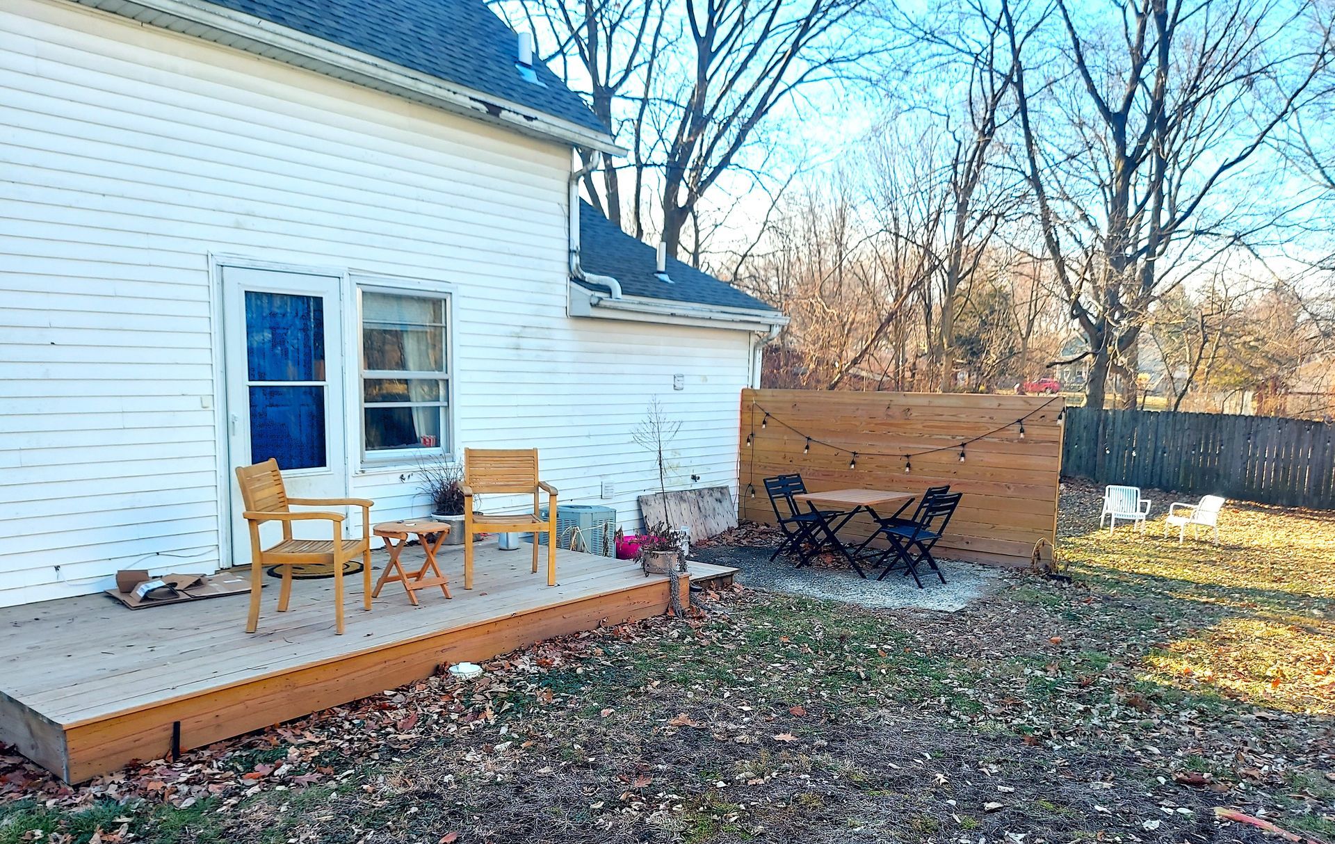 Backyard with a wooden deck, chairs, table, and a wooden privacy screen. Fall leaves on the ground.