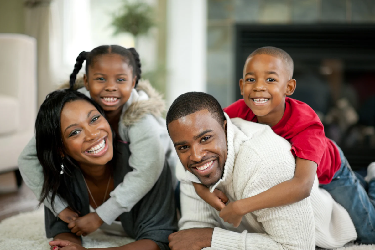 Family of four smiling and cuddling on a living room floor.