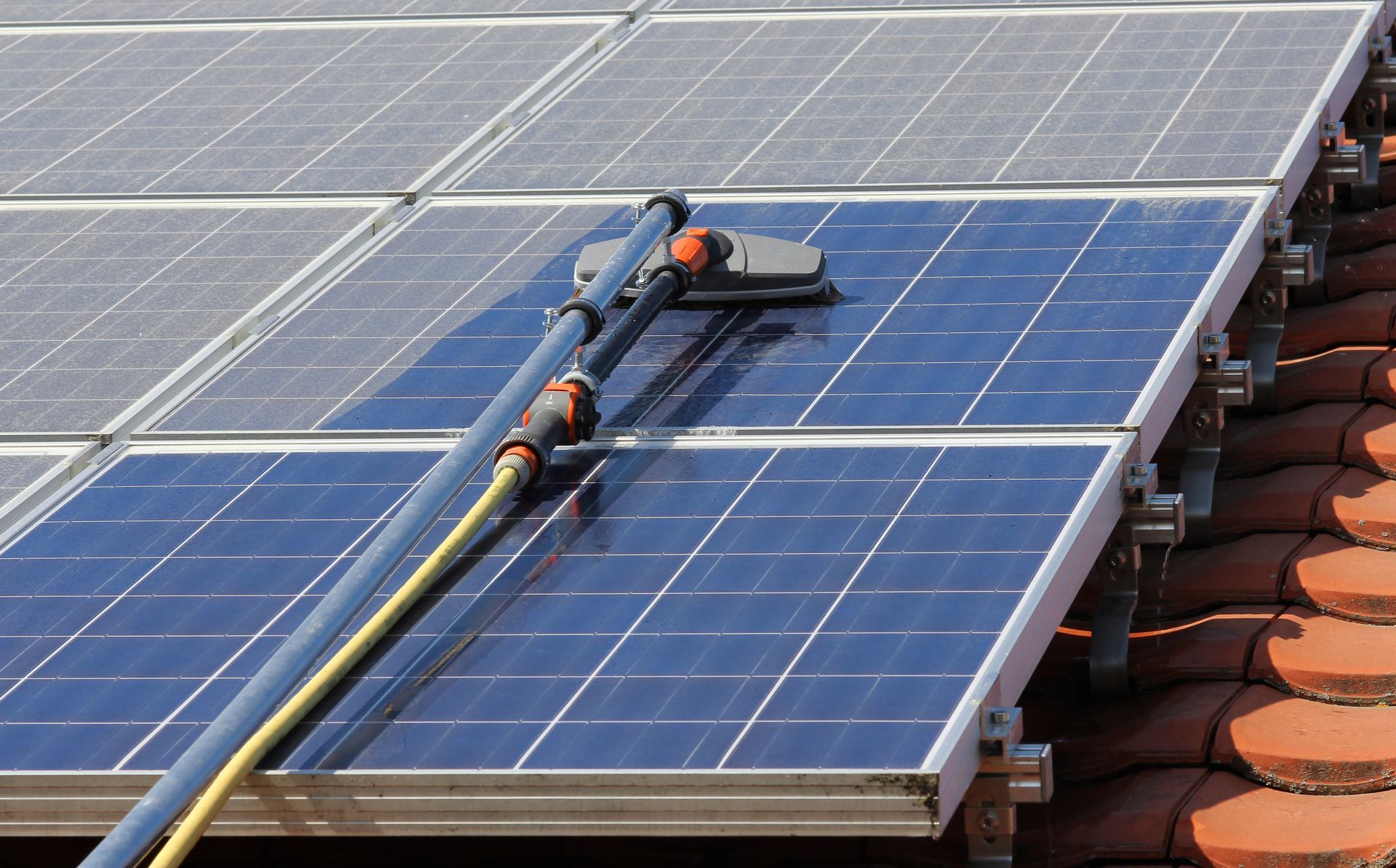 A pole-mounted cleaning tool cleans a dark solar panel on a tiled roof.