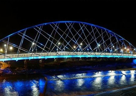 Puente arqueado iluminado de azul sobre agua oscura por la noche; las luces se reflejan en el agua.