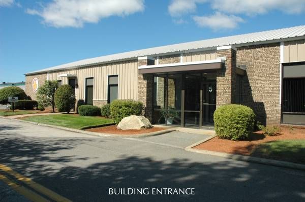 Exterior view of a beige industrial building with a glass entrance, bushes, and blue sky.