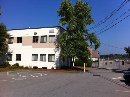 White building with brown accents, tree, and parking area. Blue sky and utility wires in the background.