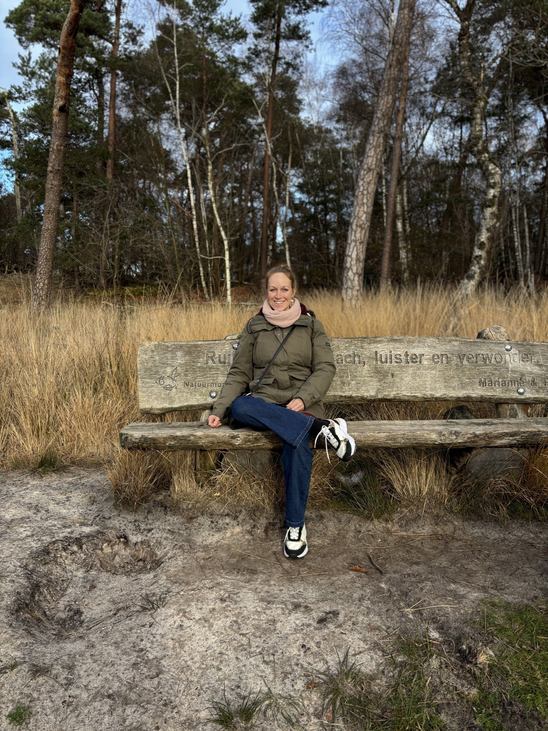 Vrouw zit op een houten bankje in een bos, gekleed in een groene jas en spijkerbroek.