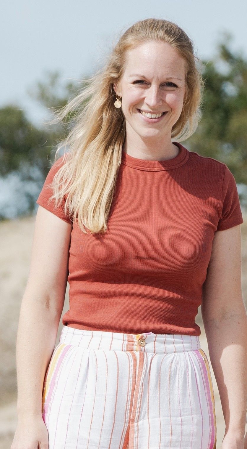 Vrouw met blond haar, gekleed in een roestbruine top en een witte gestreepte broek, lacht in de buitenlucht.