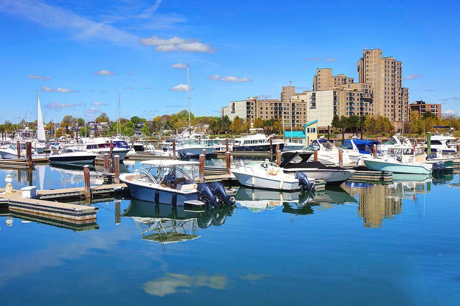 Boats docked at a marina on a sunny day with tall apartment buildings in the background under a clear blue sky.