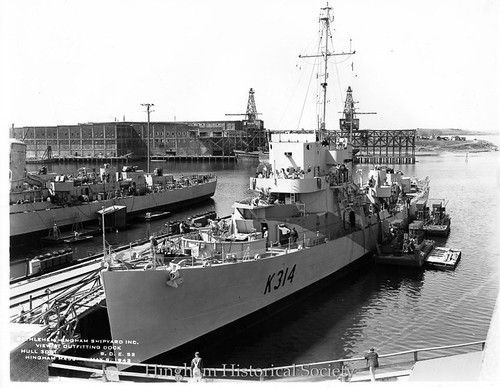 A black-and-white view of a Royal Canadian Navy frigate, K314, moored at a shipyard dock with industrial cranes behind it.