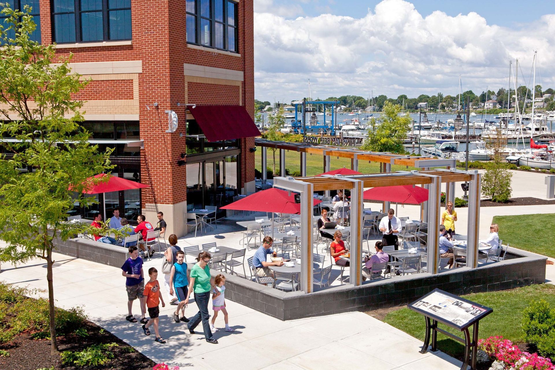 Outdoor patio seating with red umbrellas beside a brick building, overlooking a marina on a sunny day.