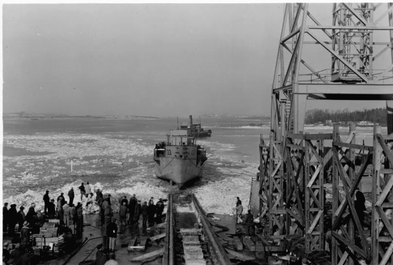A ship launching into icy water from a slipway, with a group of onlookers gathered on the left and industrial scaffolding.