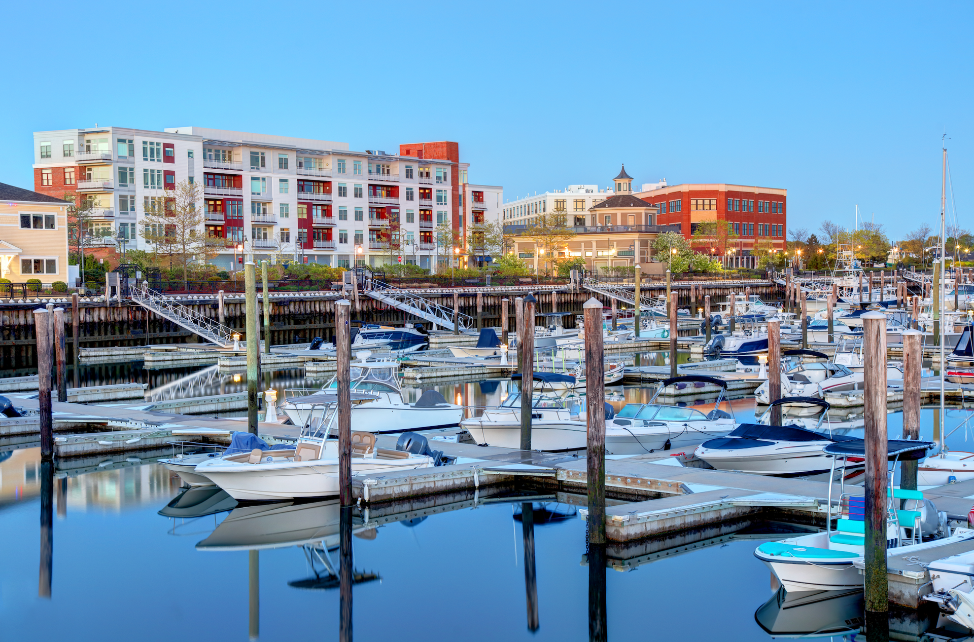 A marina filled with docked motorboats under a clear blue sky, with apartment buildings and a waterfront plaza behind.