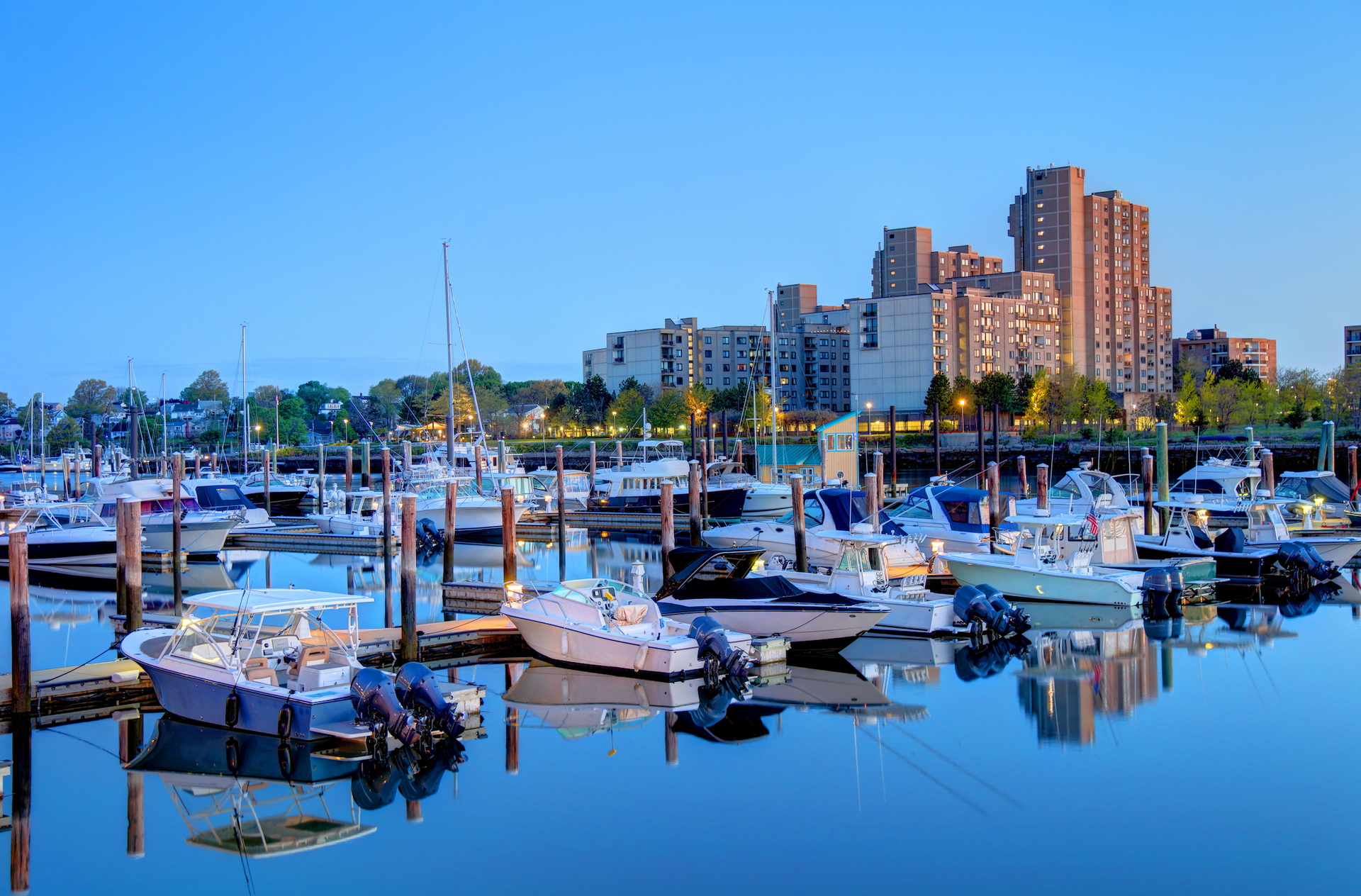 A marina with numerous small boats docked in calm water, with a background of large residential buildings at dusk.