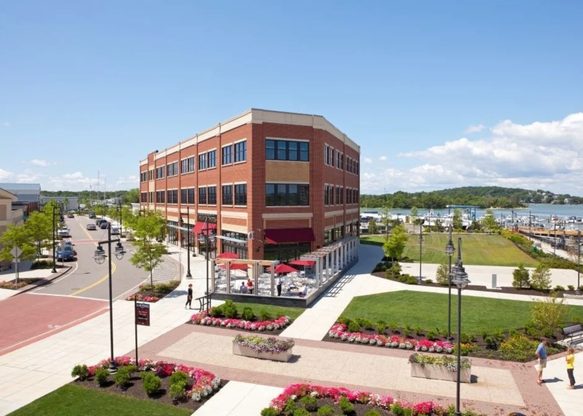 A three-story brick building with outdoor dining, adjacent to a waterfront park with walkways and manicured flower beds.