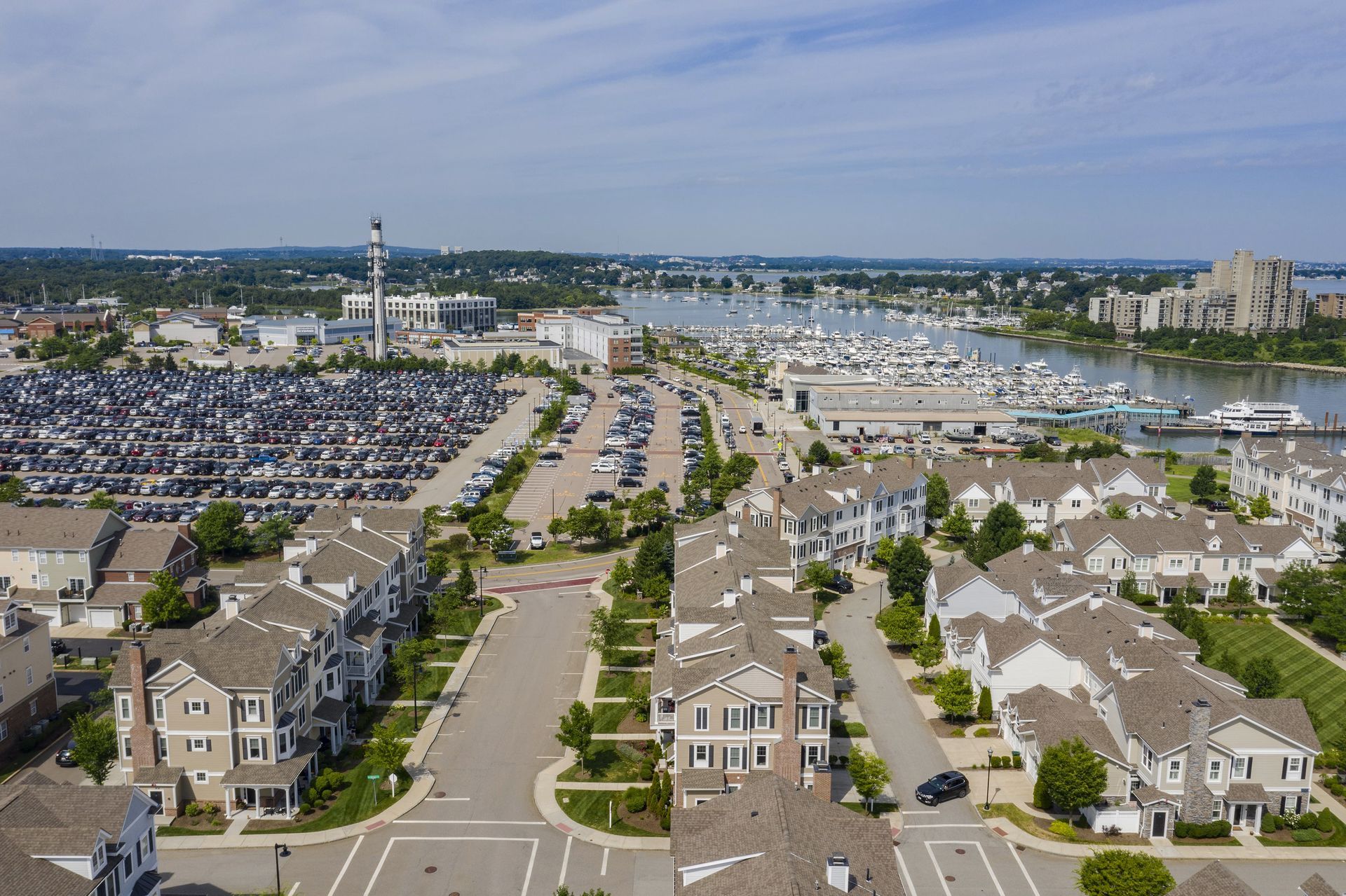 Aerial view of a coastal neighborhood with townhouses, a large parking lot, and a boat marina on a sunny day.