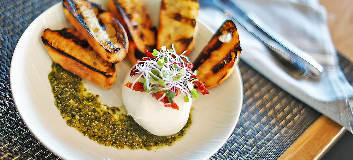 A white plate featuring grilled bread, pesto sauce, and a ball of burrata cheese topped with sprouts and red peppers.