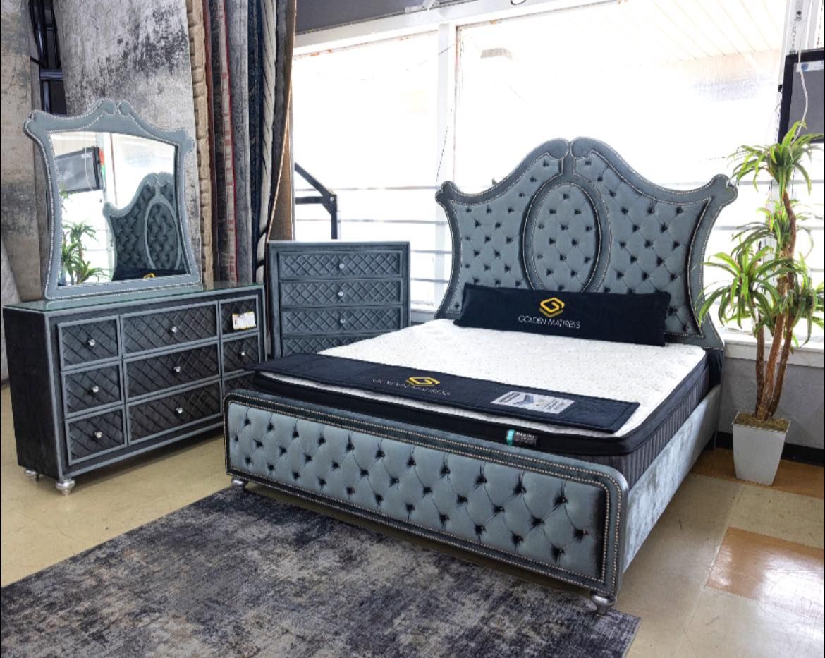 Bedroom set: gray tufted bed, dresser, mirror, chest, rug, & plants in a brightly lit room. Bayou furniture New Orleans