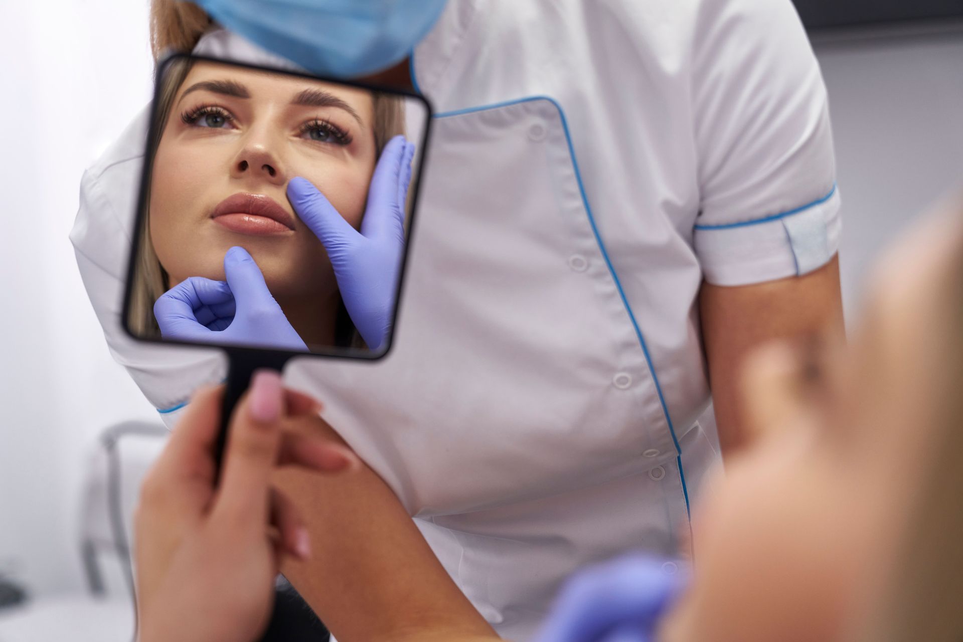 Person in blue gloves examining another person's face in a hand mirror. Clinic setting.