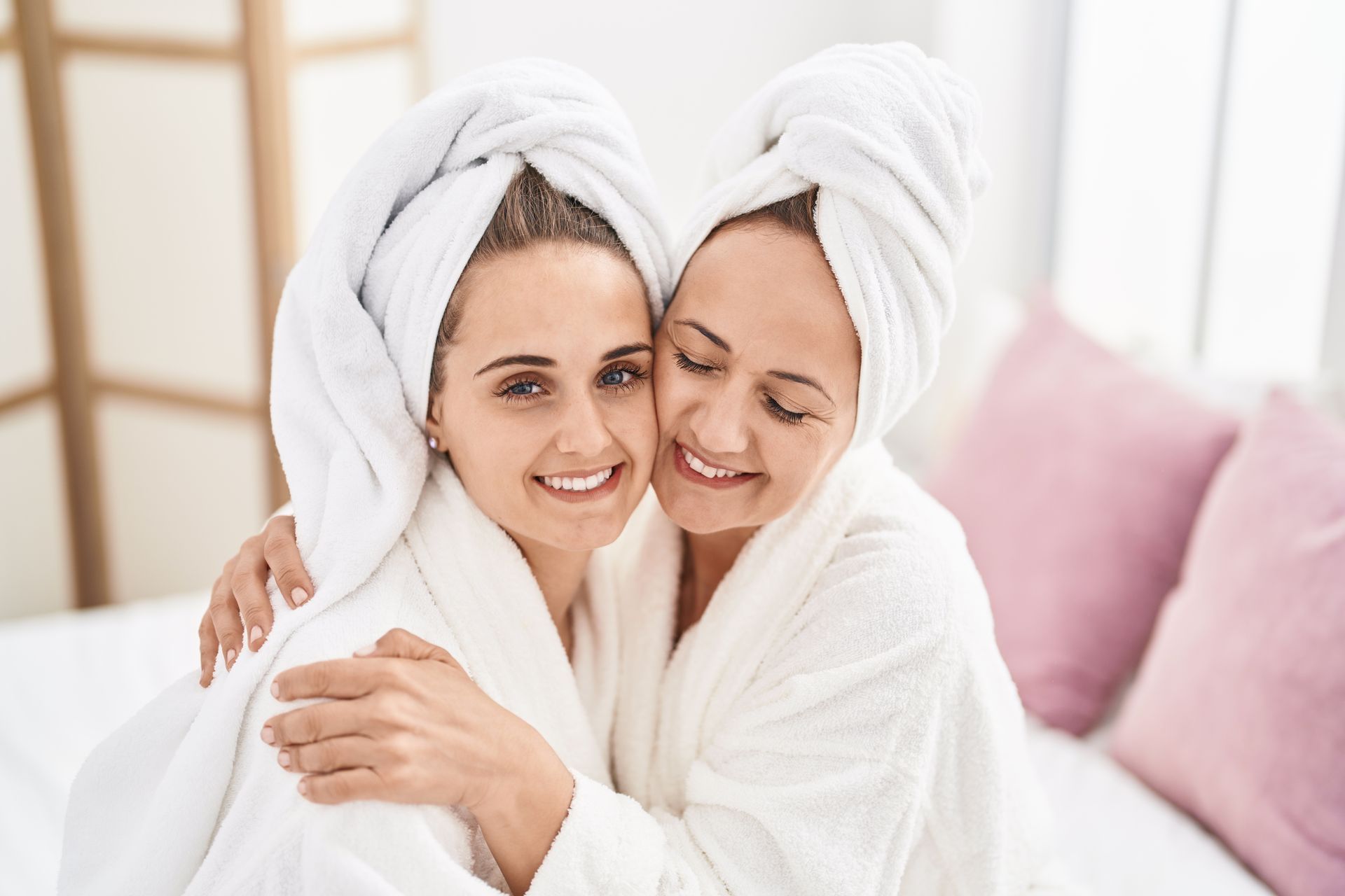 Two women in white robes and towels, hugging, smiling, indoors.