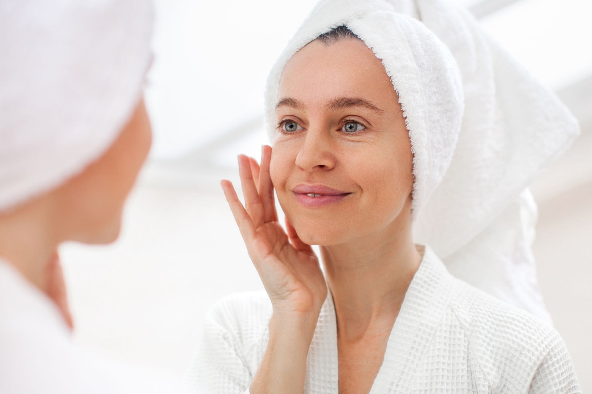 Woman in a bathrobe and towel looking in a mirror, touching her face.
