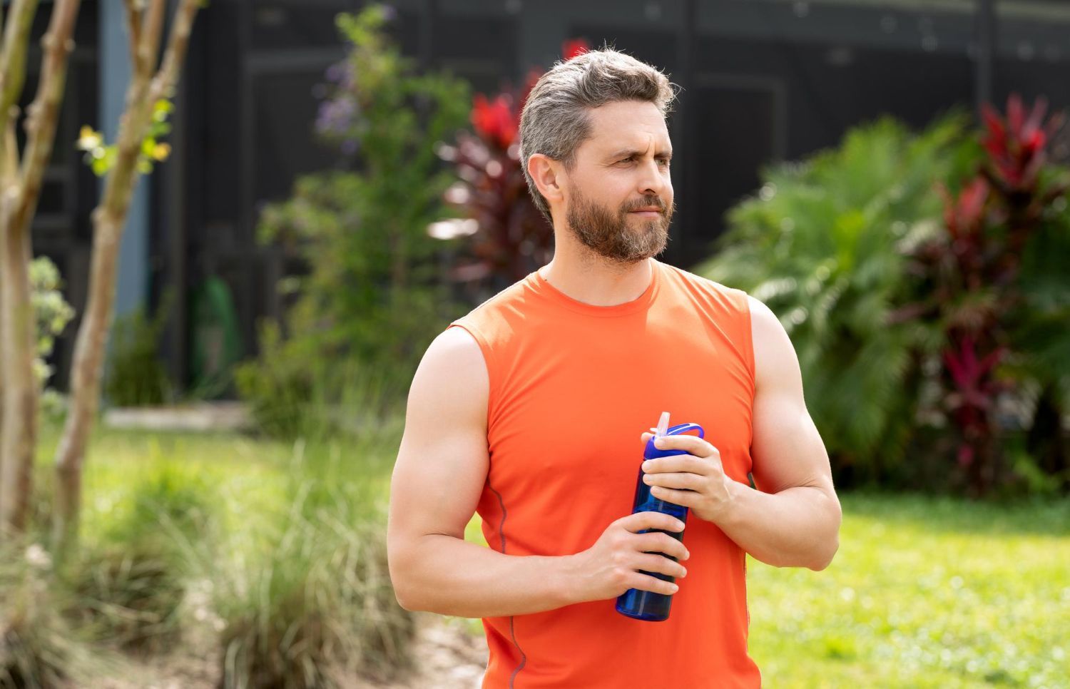 Man in orange sleeveless shirt holding a blue water bottle outdoors in a park