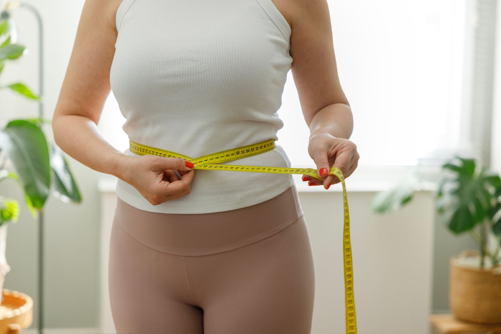 Person measuring their waist with a yellow tape measure, indoors, near plants.