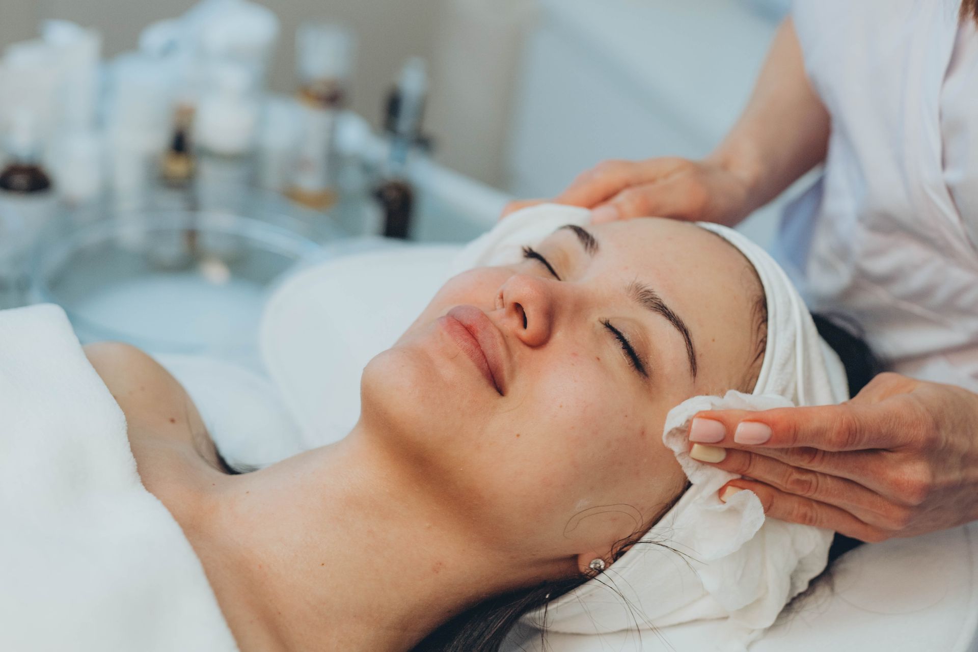 Woman receiving facial treatment; therapist cleans face with a cotton pad in a spa.