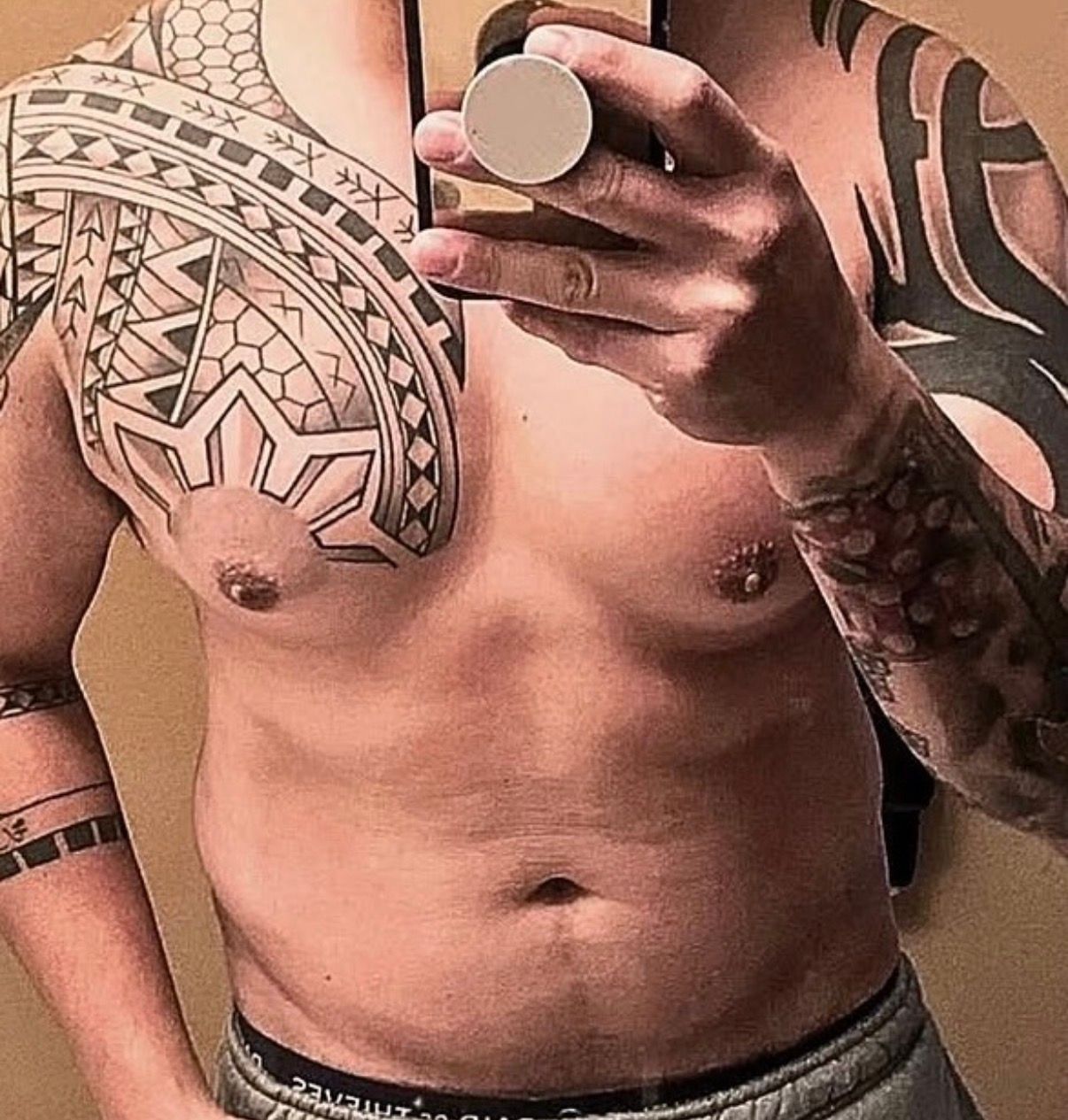 Mirror selfie of a tattooed torso in a sports bra, wearing dark shorts and holding a phone