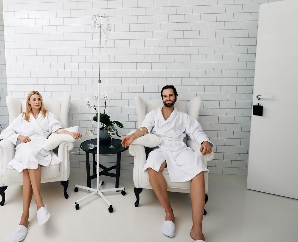 Two people in robes receiving IV drips, sitting in white armchairs in a white room.
