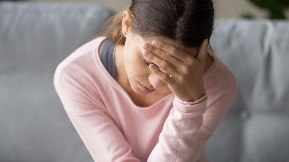 Woman sitting on a couch, holding her hand to her forehead, looking down, seemingly stressed.