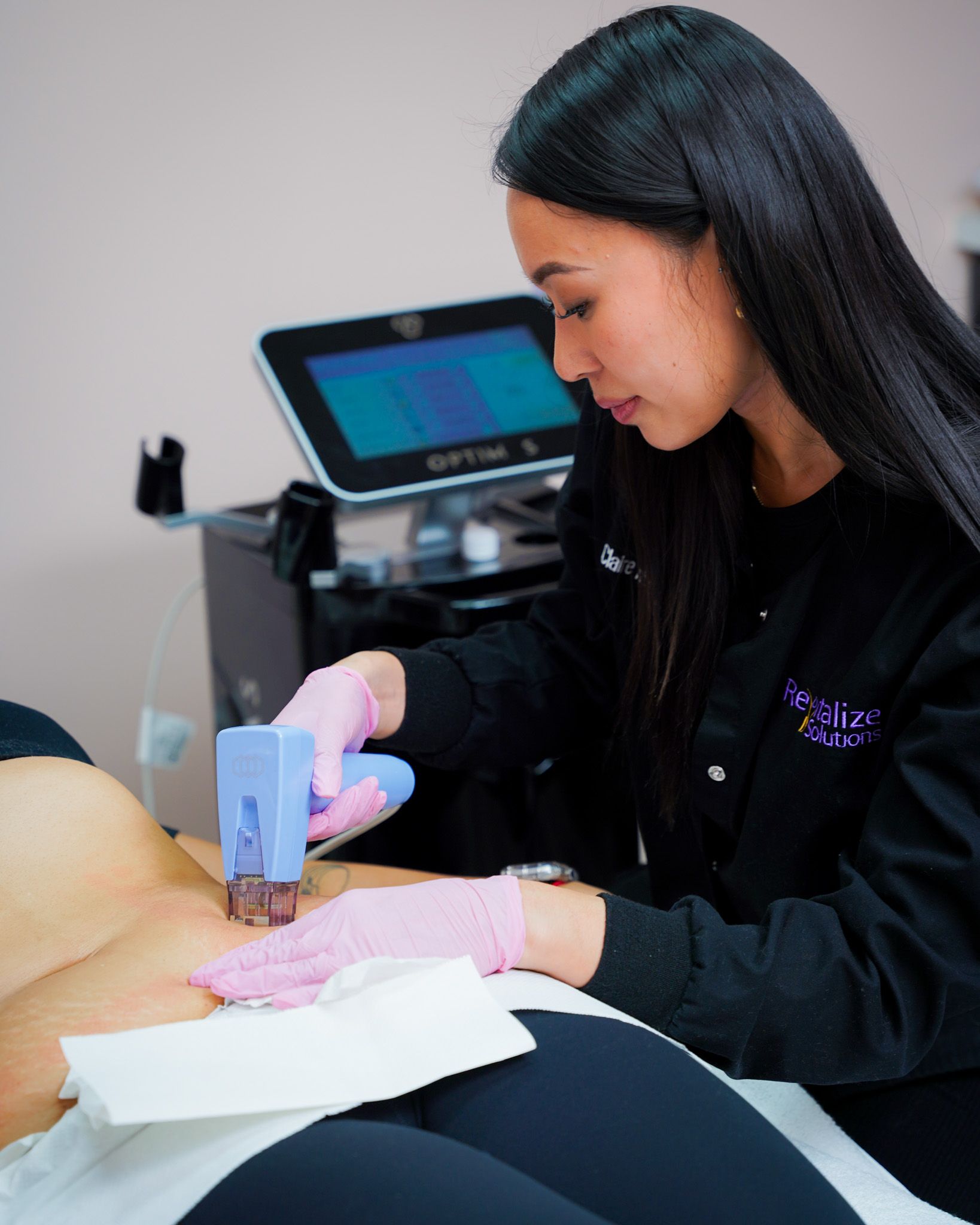 Woman performing a skin treatment, using a device on a patient's abdomen. Pink gloves and a medical setting.