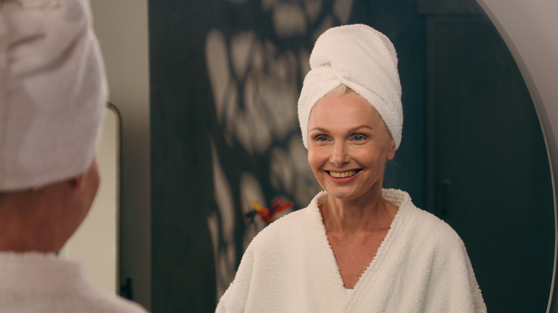 Smiling woman in a white bathrobe and towel turban, looking into a mirror in a bathroom