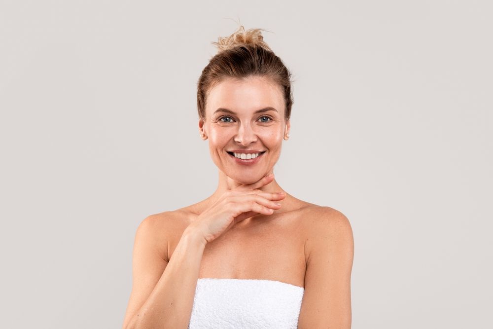 A woman wrapped in a white towel is smiling and touching her face.