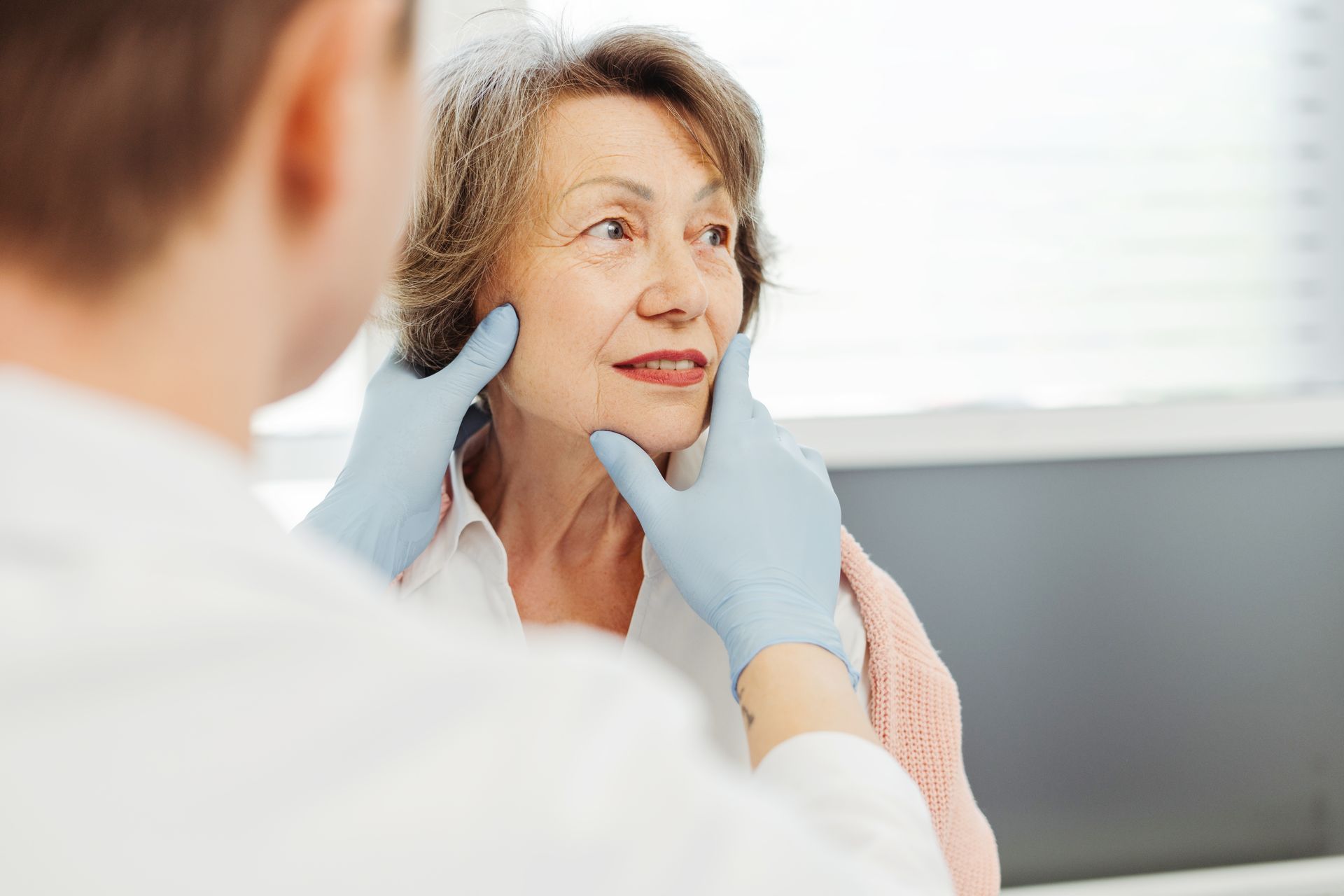 Doctor examines a patient's face with gloved hands. Patient in white shirt looks away.
