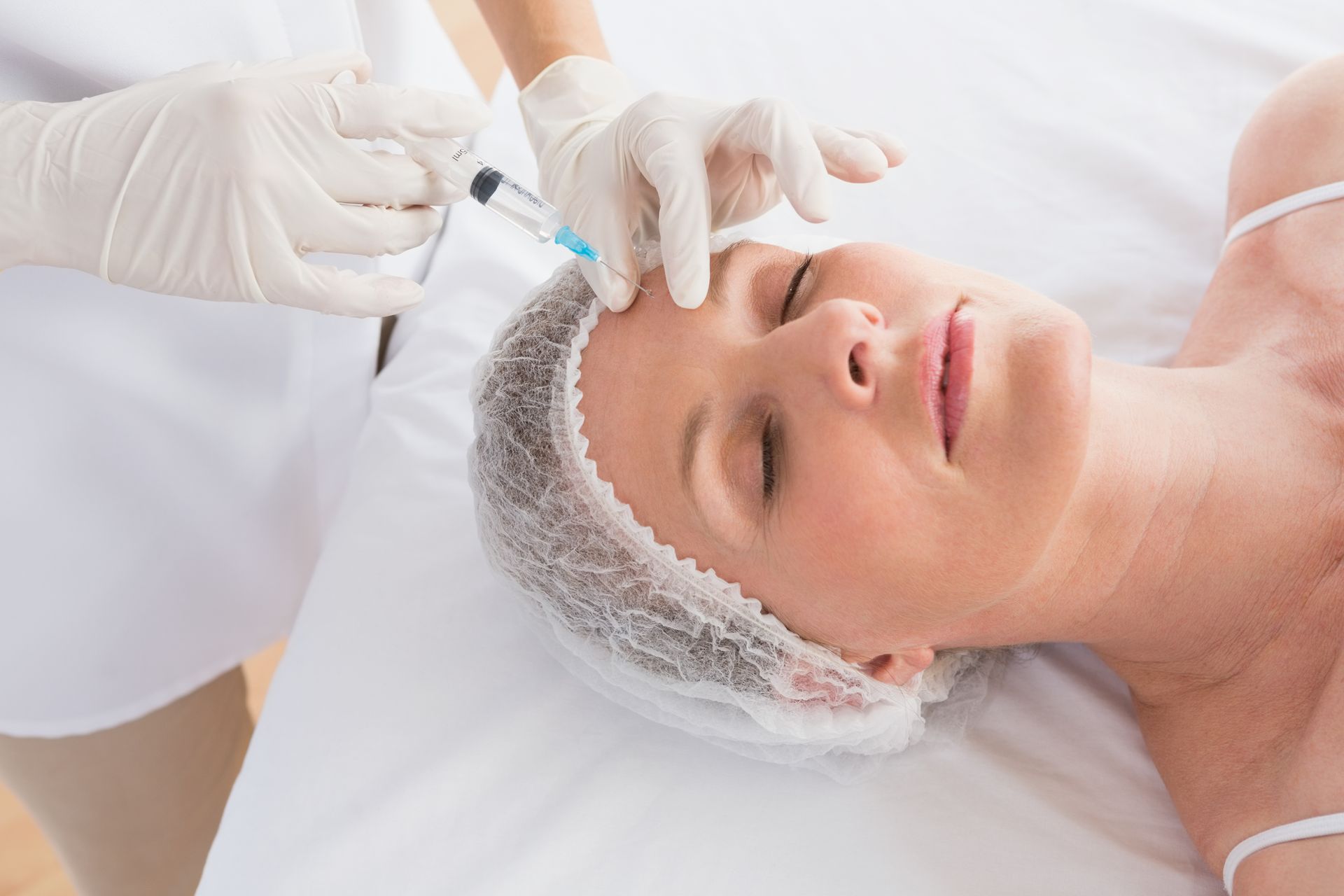 Cosmetologist injecting facial treatment into a woman’s forehead with a syringe, wearing gloves and a hair cap