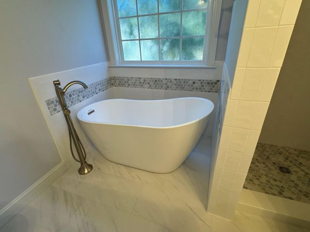 White freestanding bathtub near a window in a bathroom.  Brass faucet, white and gray tile.