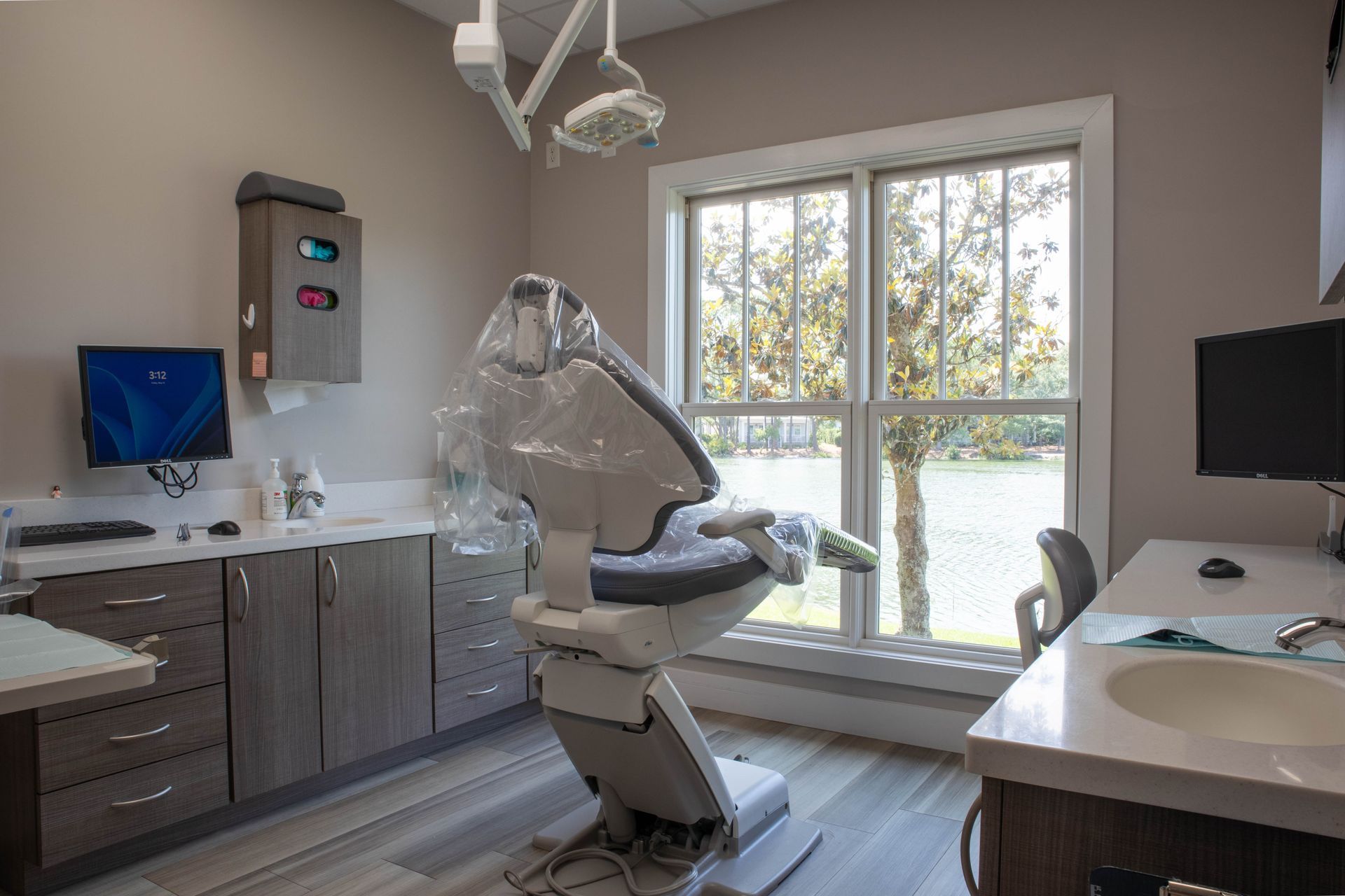 Dental exam room with chair, cabinets, monitors, and large window overlooking greenery