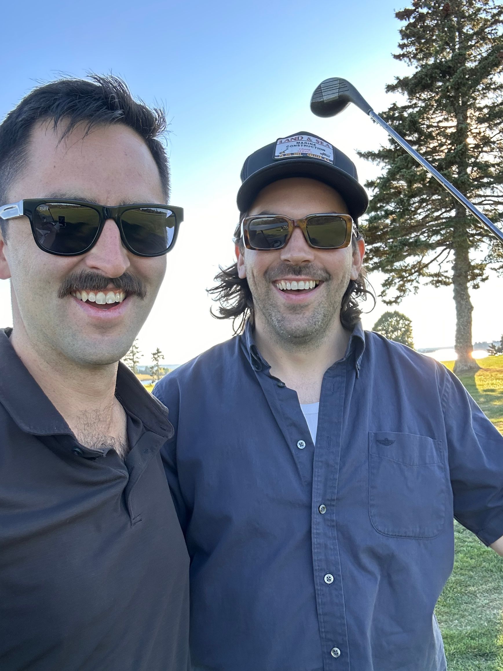 Two smiling people outdoors in sunglasses, one holding a golf club at sunset.