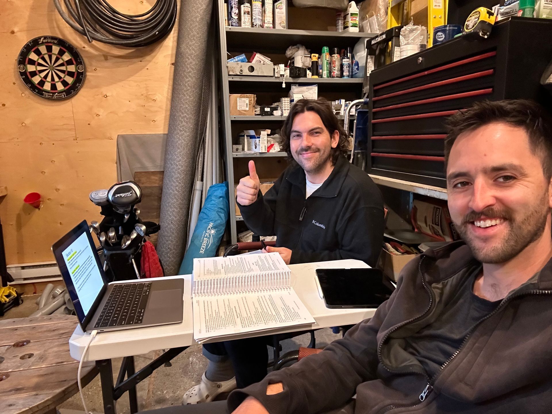 Two people smiling in a workshop, seated at a table with a laptop, whiteboard, and tools.