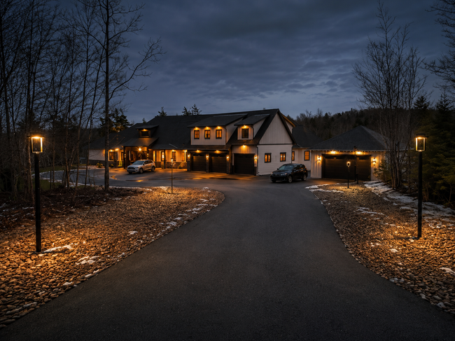 Large house at dusk with warm exterior lights, long driveway, and bare trees under a cloudy sky