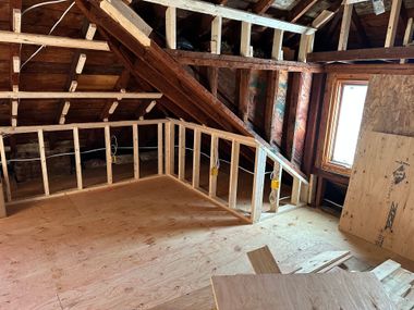 Unfinished attic interior with exposed wooden framing, sloped rafters, and a bright window.