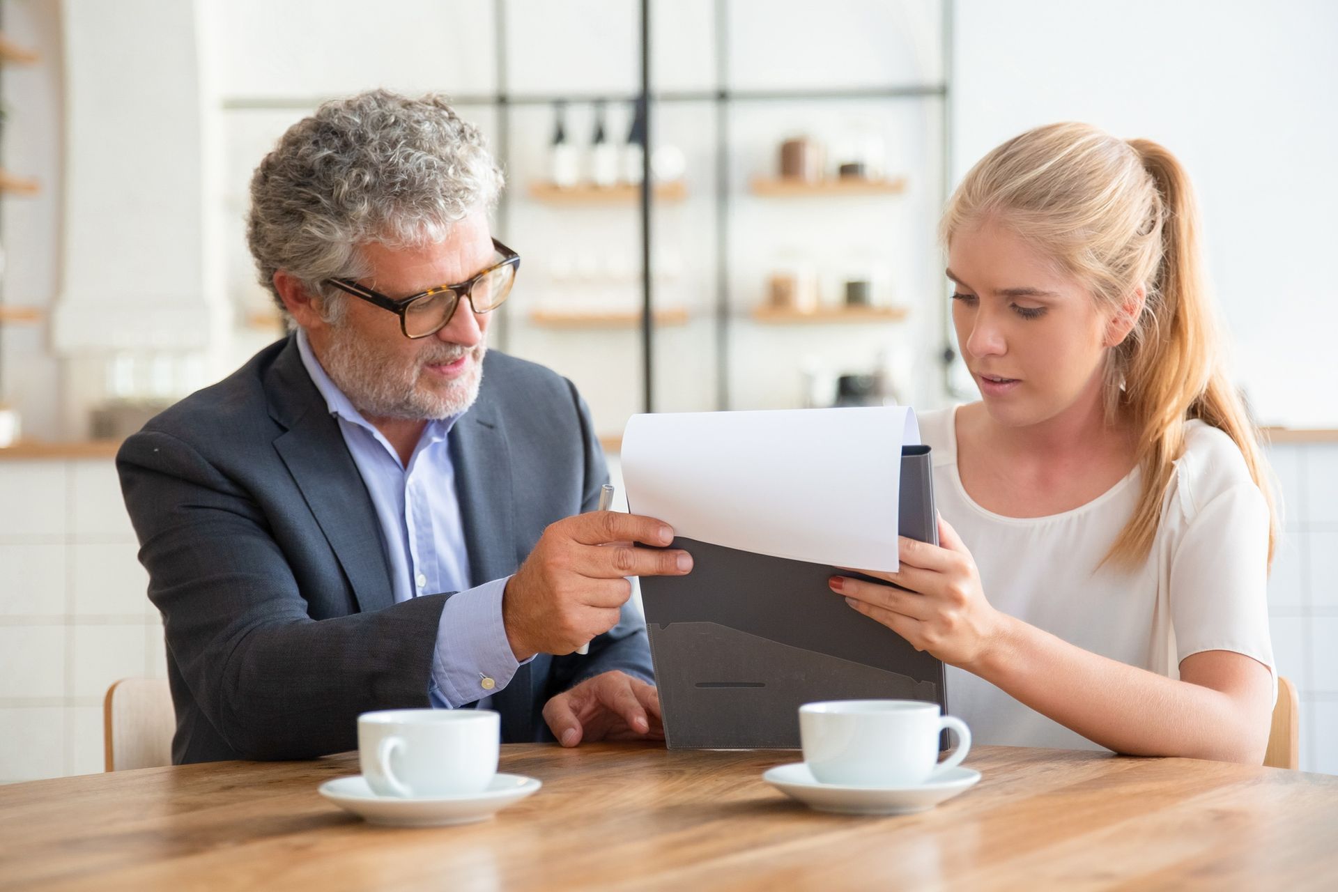 A man in a suit and a woman are reviewing documents at a table. Two coffee cups are present.