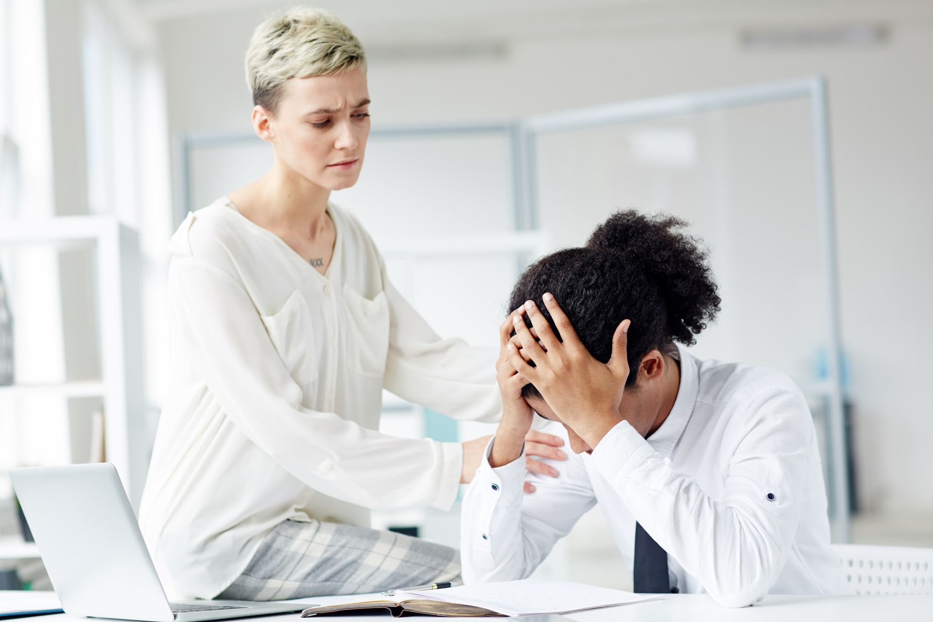 A woman is comforting a man who is sitting at a desk with his head in his hands.