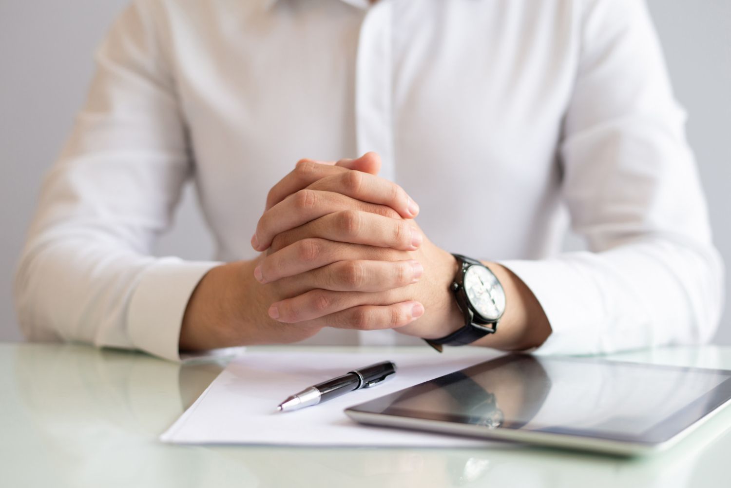 Hands clasped on a table with a pen, paper, and tablet, person in a white shirt.