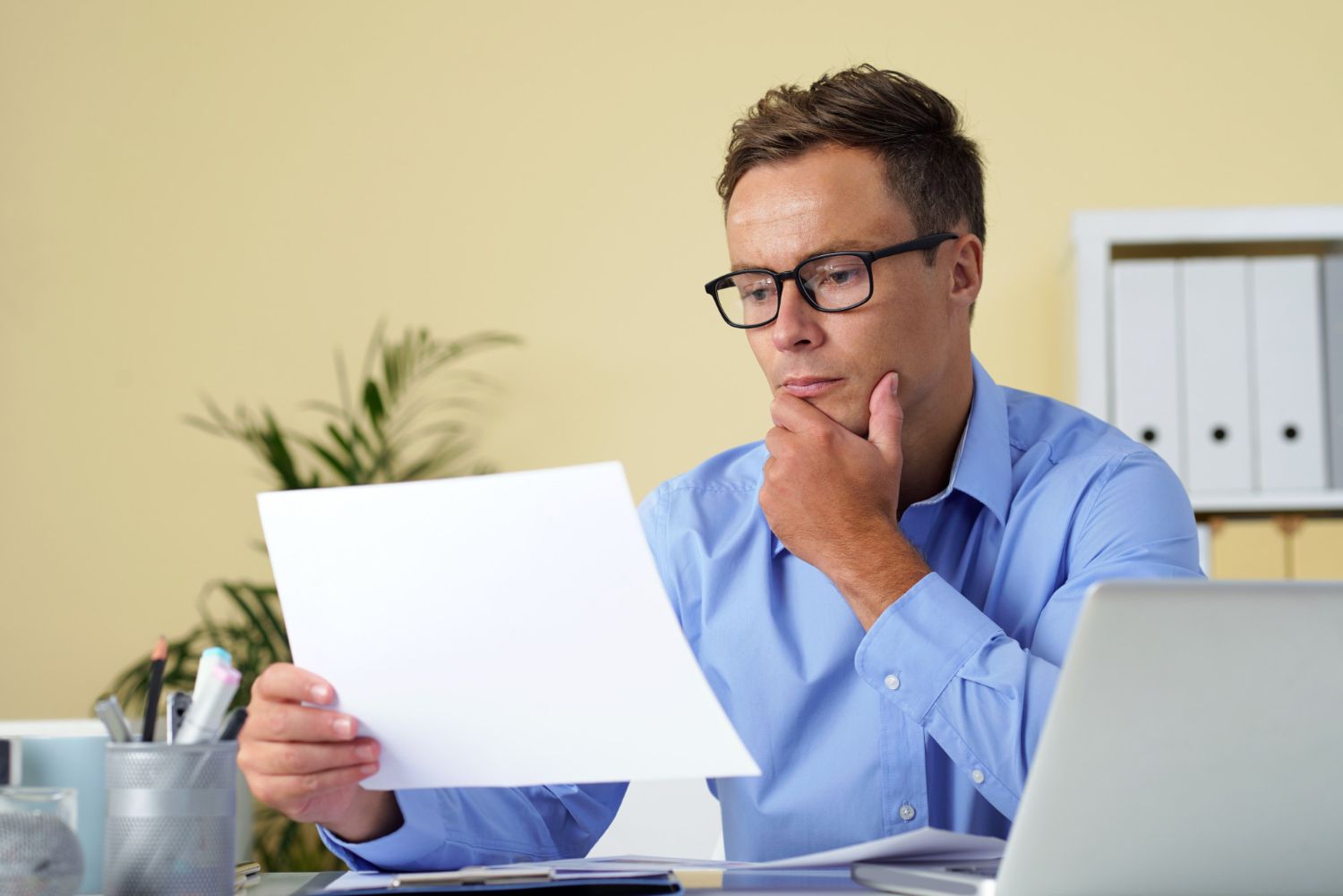 Man with glasses, blue shirt, at desk reviewing paperwork, thoughtful expression. Office setting, laptop visible.
